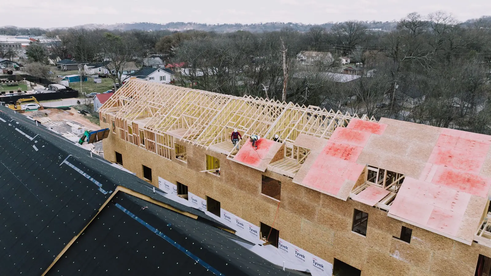Drone view down the roofline of a long wood-framed apartment building mid-sheathing, with the working site around it.